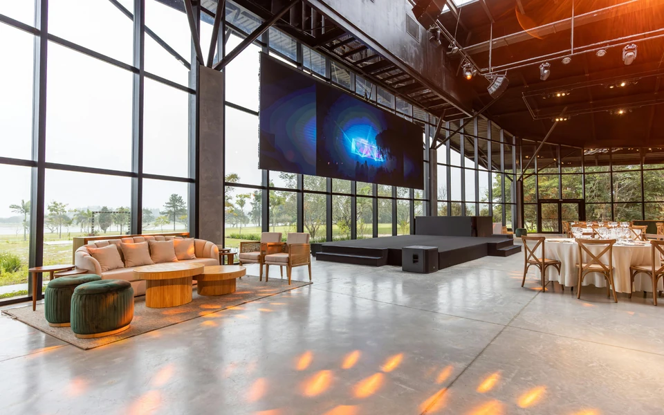 Espaço para eventos com palco, ecrã, zona de estar e mesas preparadas.